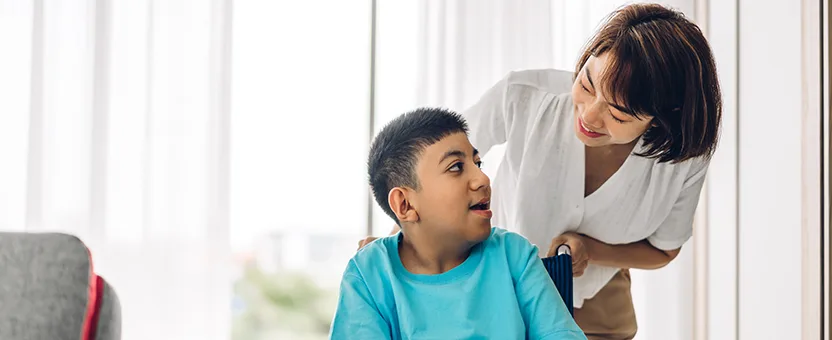 A nurse stands behind a young man with disabilities in a wheelchair. She is leaning down and smiling at him.